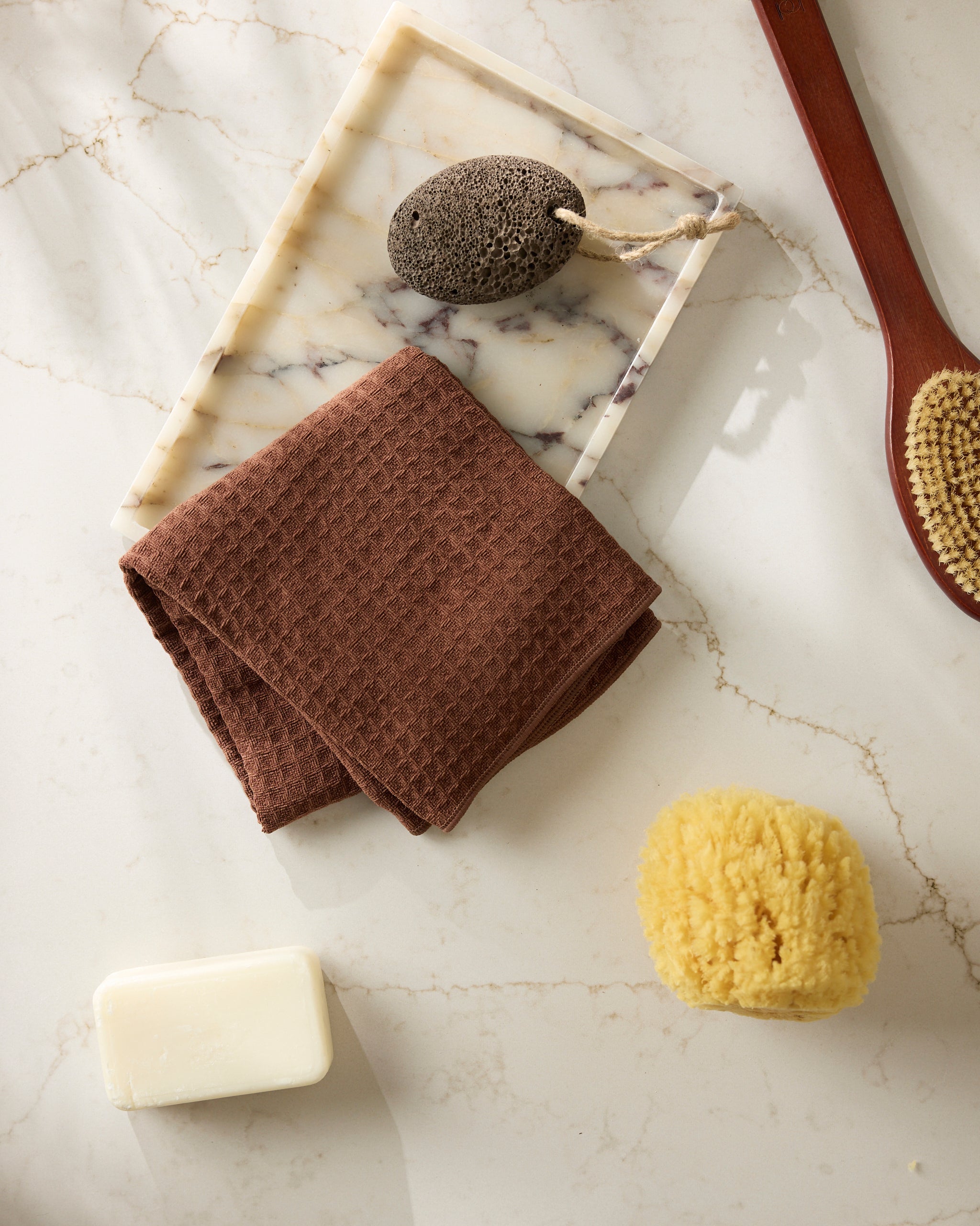 Geometry’s Maple textured towel, a pumice stone, bath brush, yellow sponge, and white bar of soap are arranged on a white marble surface alongside a marble tray.