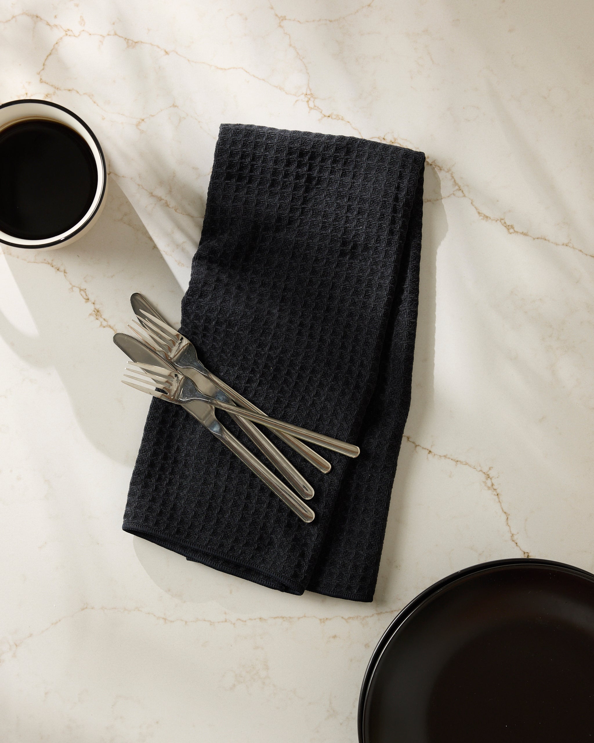 The Geometry Onyx napkin, featuring a black textured design, holds three forks and three knives beside a coffee cup and dark plate on a light marble surface.