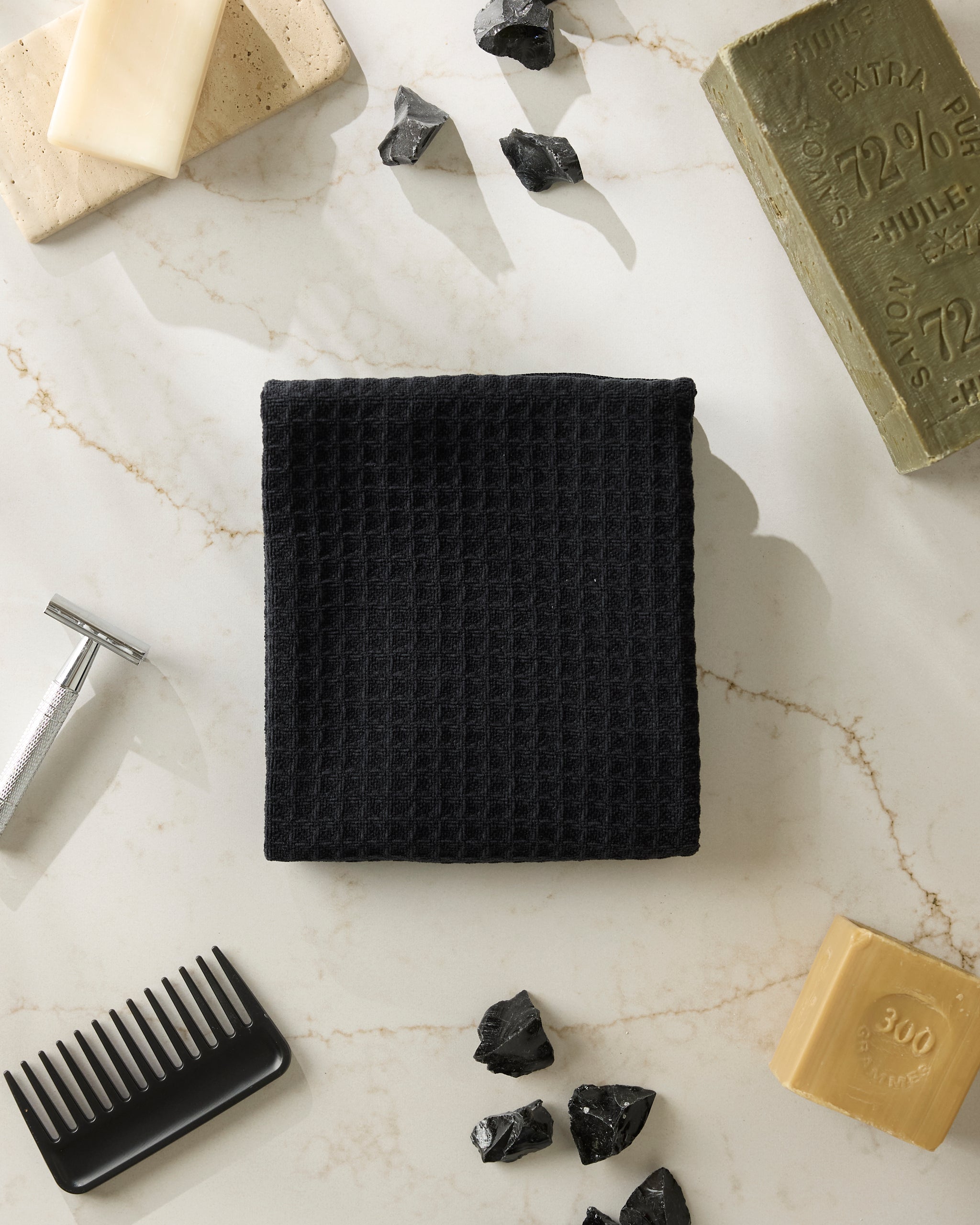 The Geometry Onyx black textured towel is centered on a marble surface, surrounded by assorted soaps, black stones, a metal razor, and a black comb.