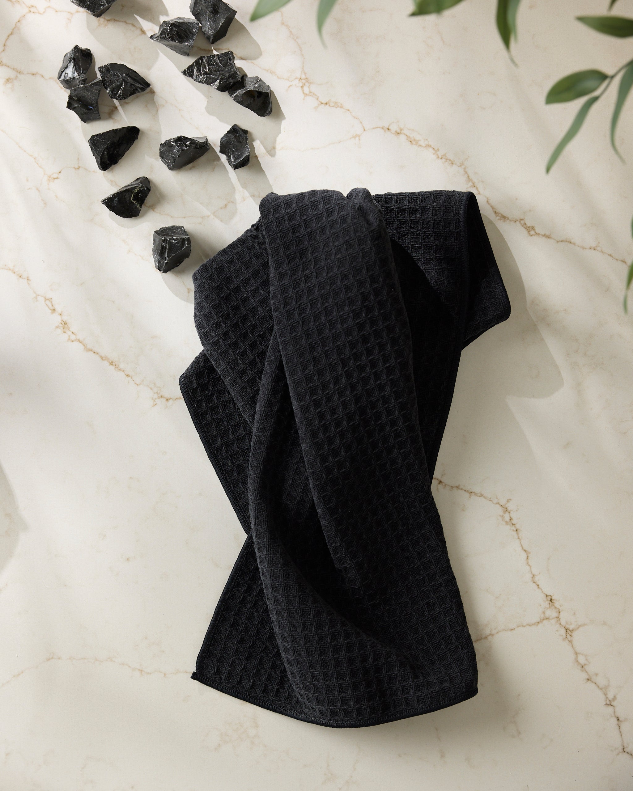 The Onyx towel by Geometry is displayed on a white marble surface with beige veins. Black stones accent the upper left corner, while green leaves partially frame the top right, highlighting the towel’s textured black design.