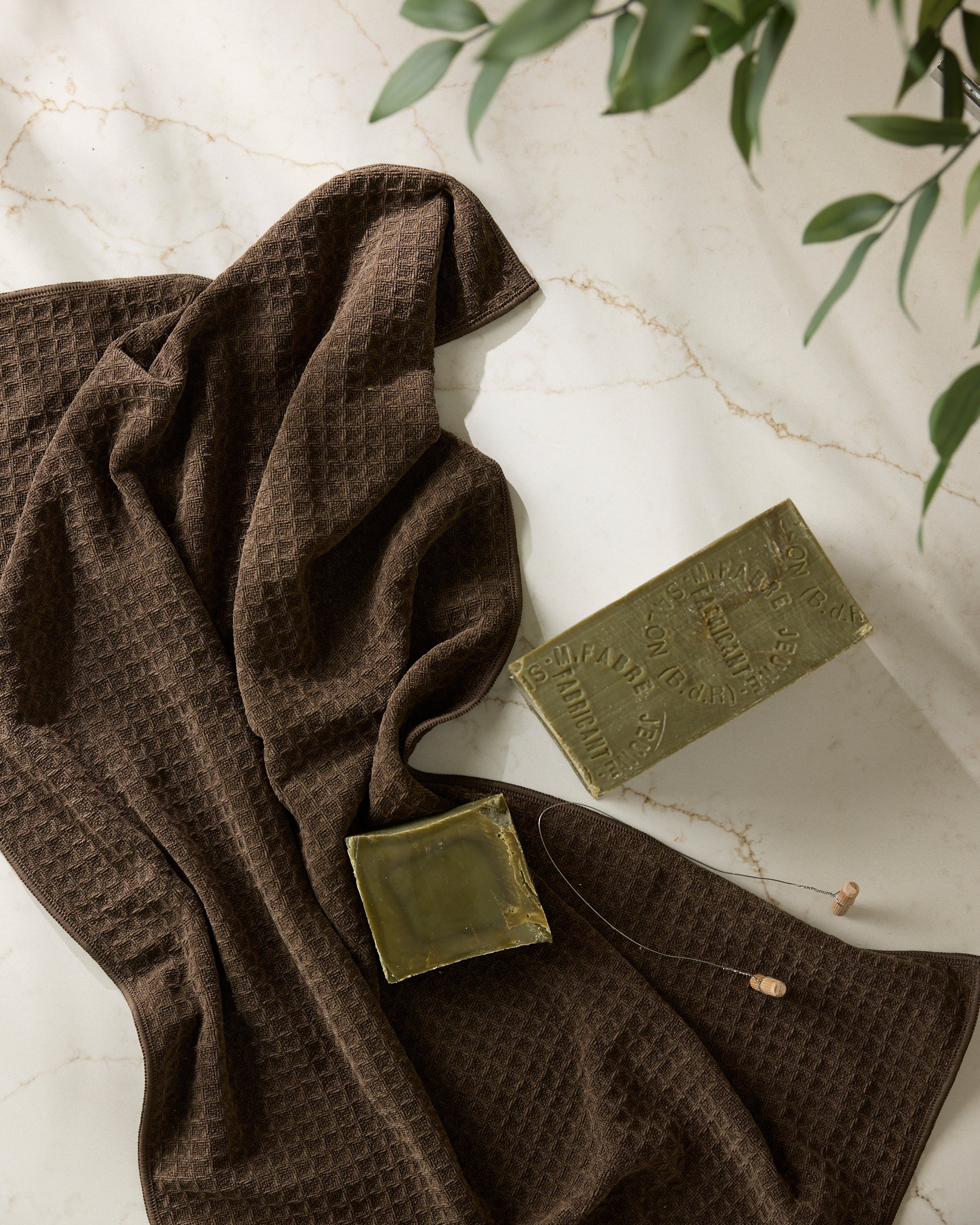 A Geometry Espresso towel lies on a marble surface beside a green soap bar, a small green dish, and a brown earplug, with green plant leaves in the corner.