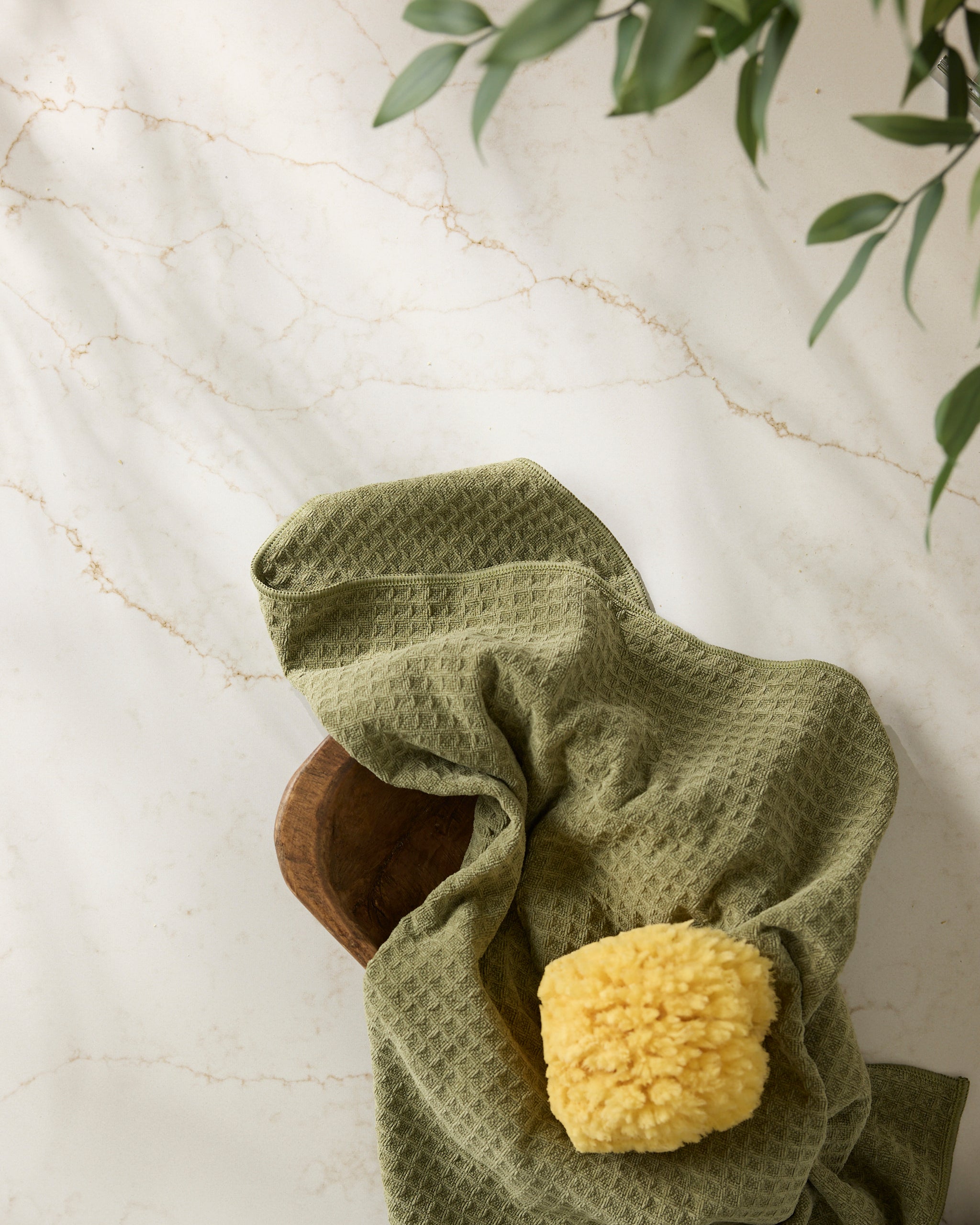 A yellow Geometry 'Olive' bath sponge sits on a textured green towel atop a wooden dish on white marble with beige veins, while green leaves hang in the upper right corner.