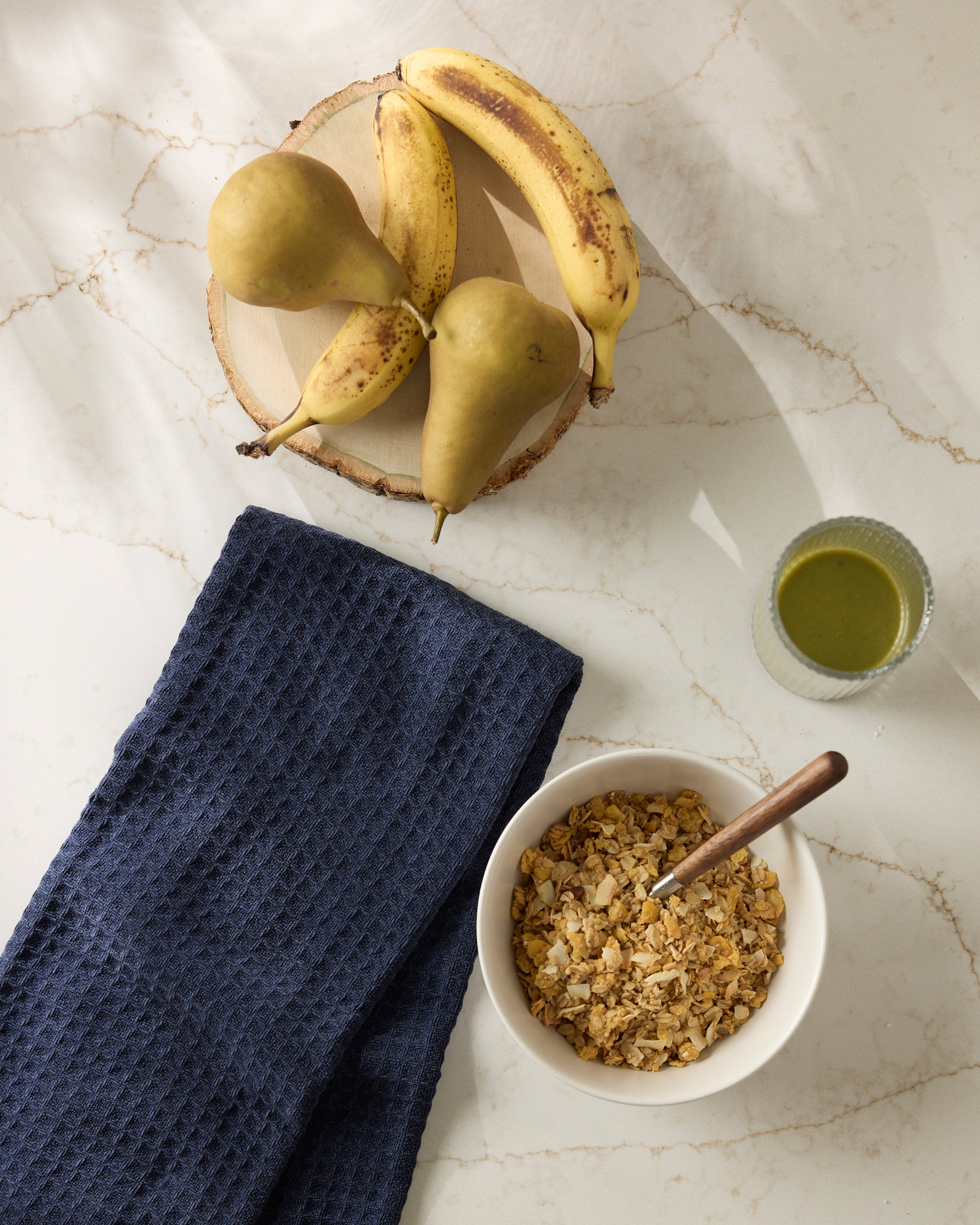 A bowl of Geometry's Sapphire granola with a spoon sits on a marble countertop next to a folded navy blue cloth, three pears and two bananas on a wooden plate, and a glass of green juice.