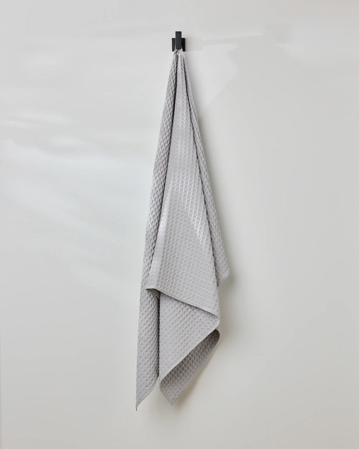 The GEOMETRY Stone waffle bath towel in light gray hangs neatly on a black hook against a plain white wall, adding style and comfort.