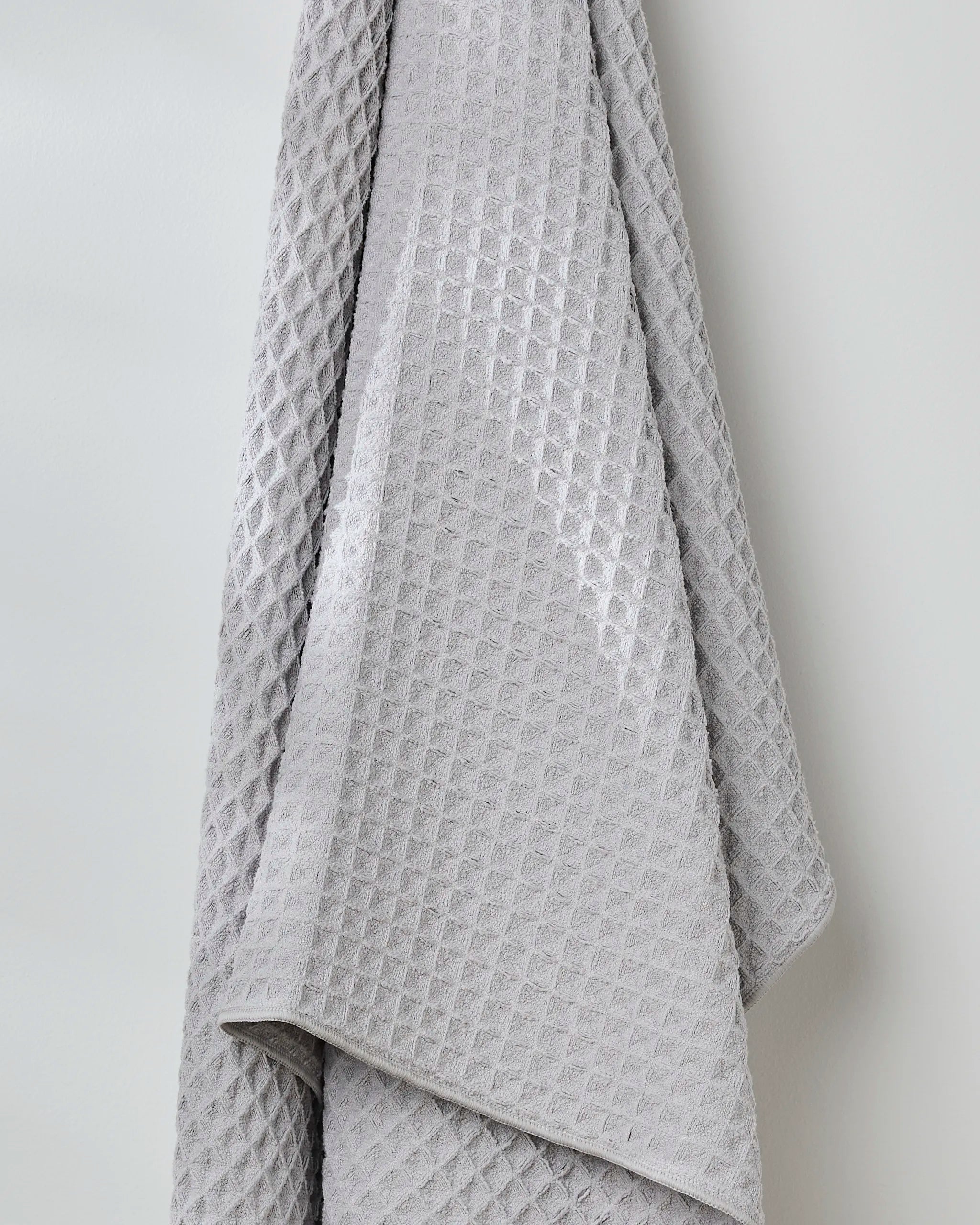 The Stone waffle bath towel by GEOMETRY, in gray with a grid-like texture, hangs in soft folds on a light wall, bringing style and comfort to your space.
