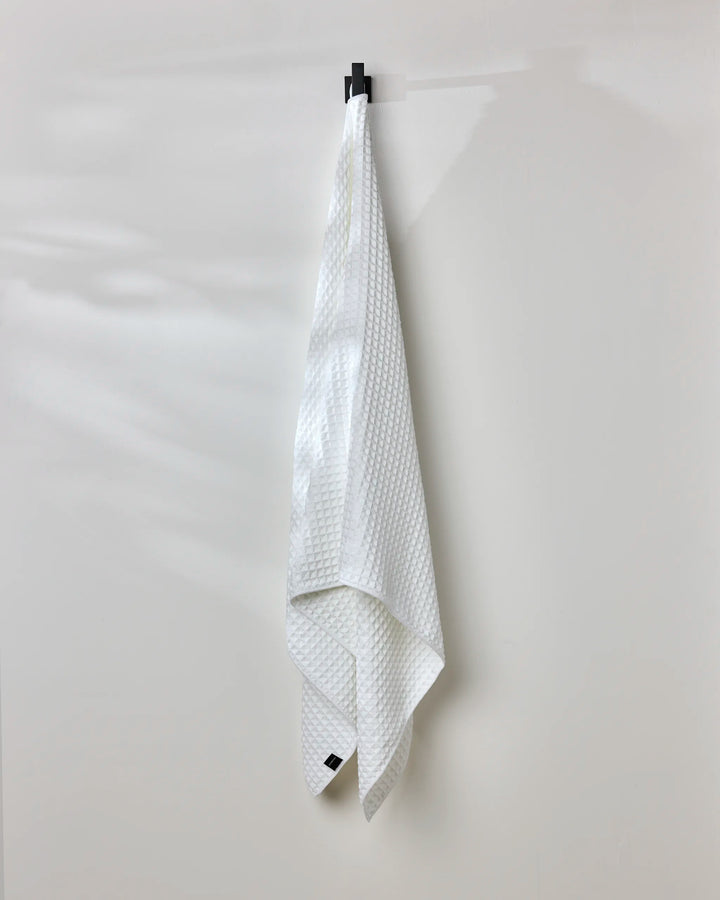 A GEOMETRY Cloud white waffle bath towel hangs neatly from a black hook on a plain, light-colored wall.