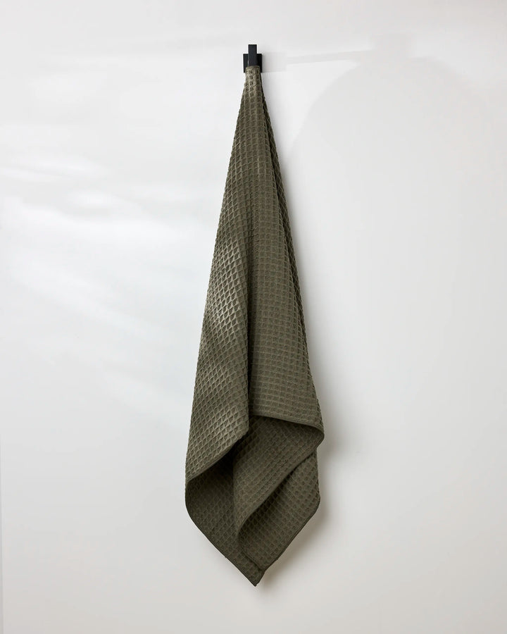 The GEOMETRY Forest bath towel in olive green, featuring a textured waffle pattern, hangs neatly on a black hook against a plain white wall, with soft folds and gentle shadows accentuating its design.