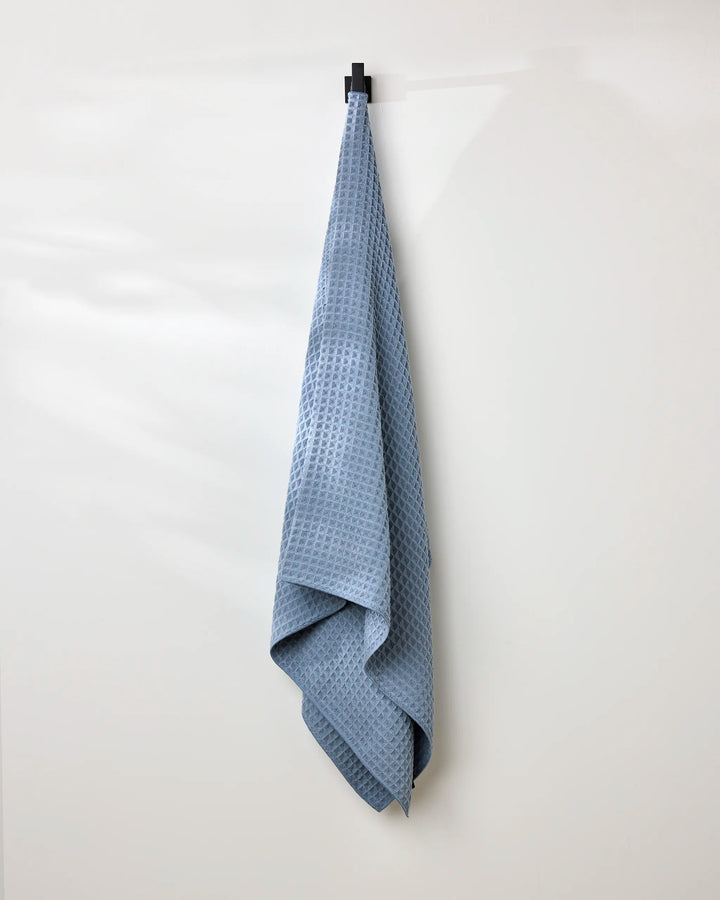 The GEOMETRY Sky waffle bath towel, in light blue, hangs neatly from a black hook on a white wall, bringing an elegant touch of simple luxury to the space.