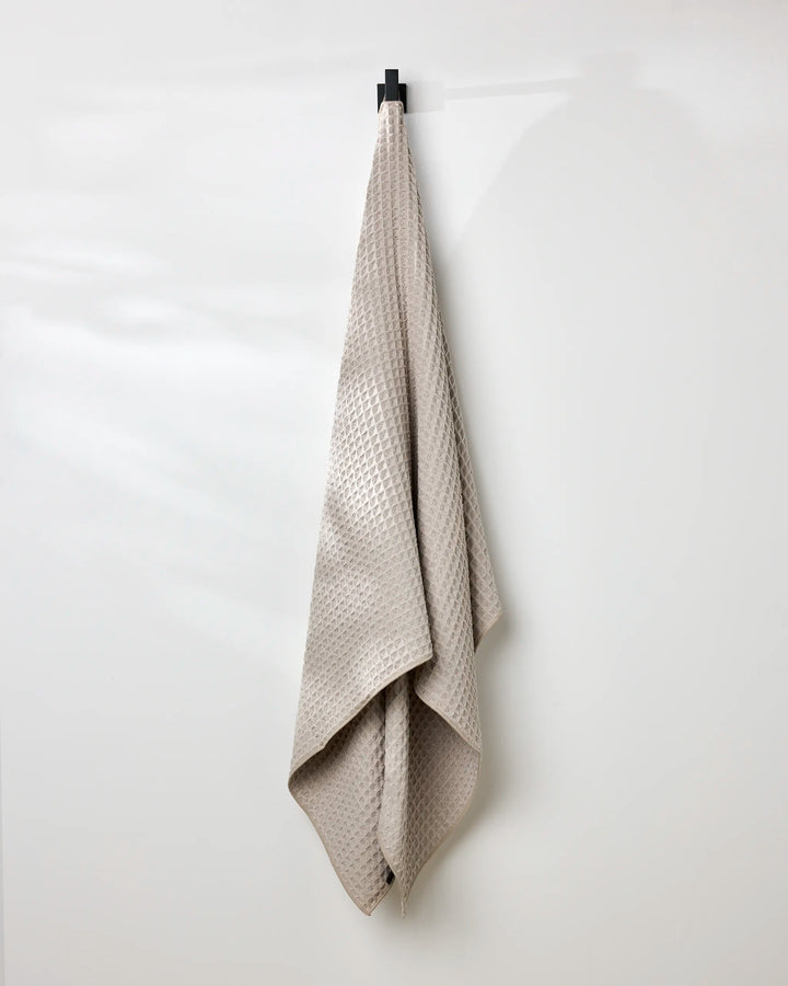 A taupe, eco-friendly GEOMETRY waffle bath towel with a textured pattern hangs neatly on a single black hook against a smooth white wall.
