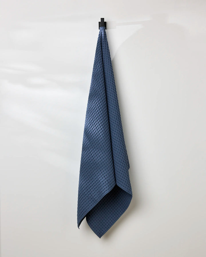 A GEOMETRY Midnight Blue bath towel hangs neatly from a black hook on a white wall, with soft light casting gentle shadows.