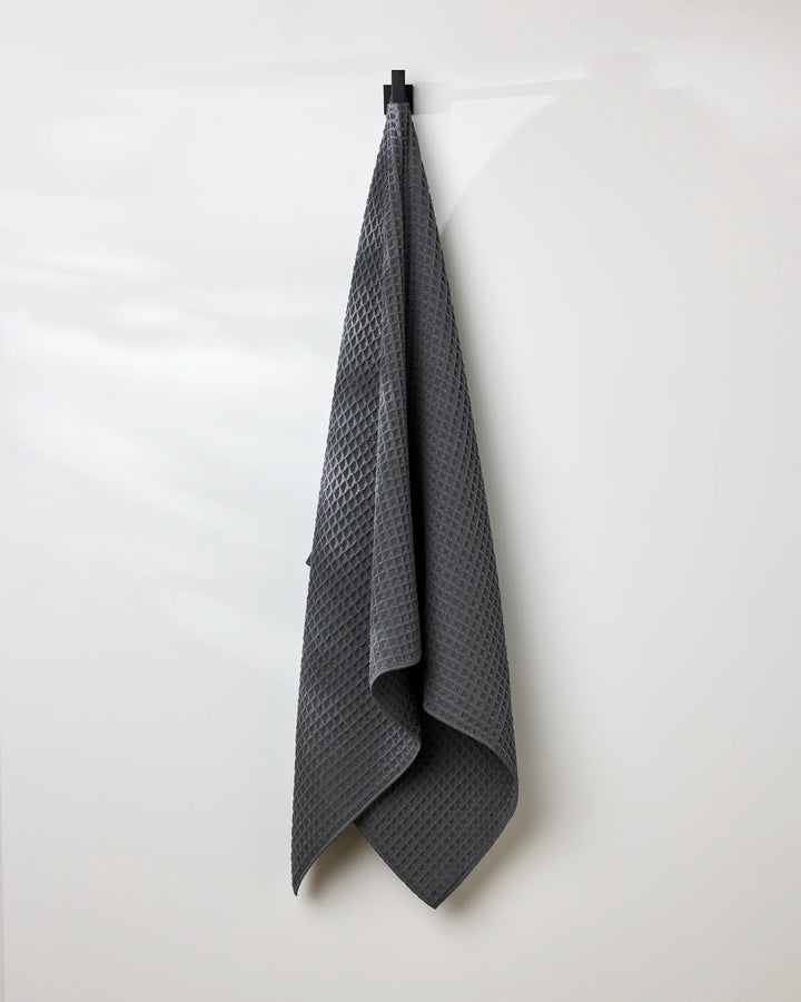 A GEOMETRY Charcoal waffle bath towel hangs neatly on a black hook against a plain white wall.
