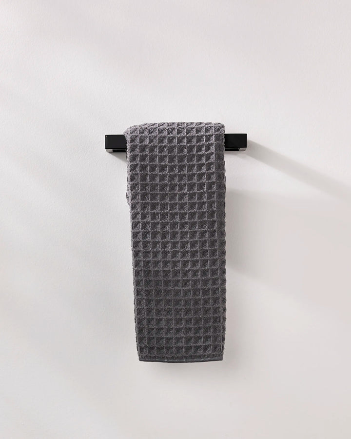 A GEOMETRY Charcoal waffle hand towel hangs neatly on a black horizontal bar against a plain white wall.