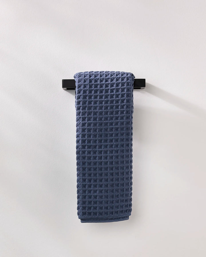 The GEOMETRY Midnight Blue luxury hand towel with a waffle texture is neatly folded over a sleek black towel bar on a white wall.