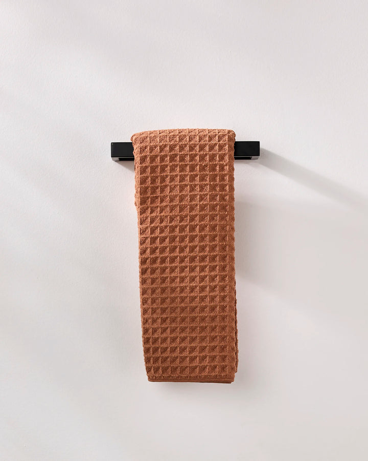 A GEOMETRY Clay waffle hand towel in a rich rust hue hangs on a sleek black bar against a white wall, bringing an eco-friendly touch to the space.