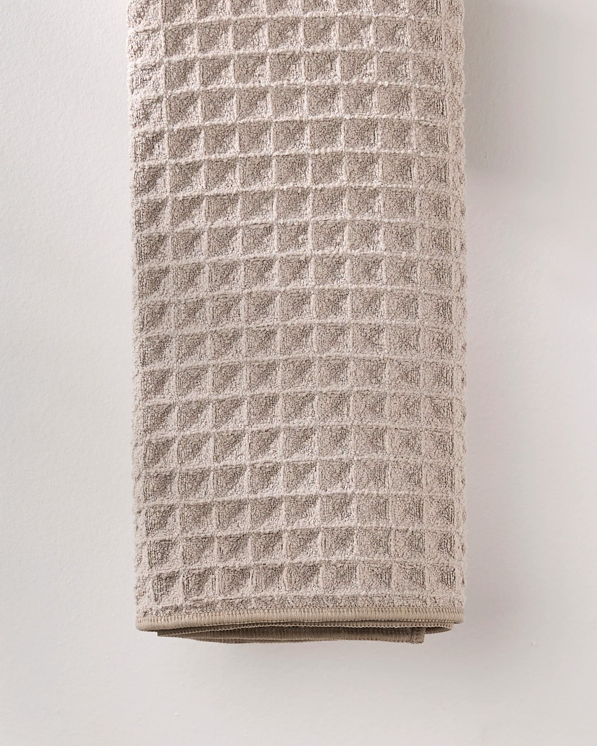 A folded GEOMETRY Taupe waffle hand towel, crafted from recycled materials, hangs against a plain white wall.