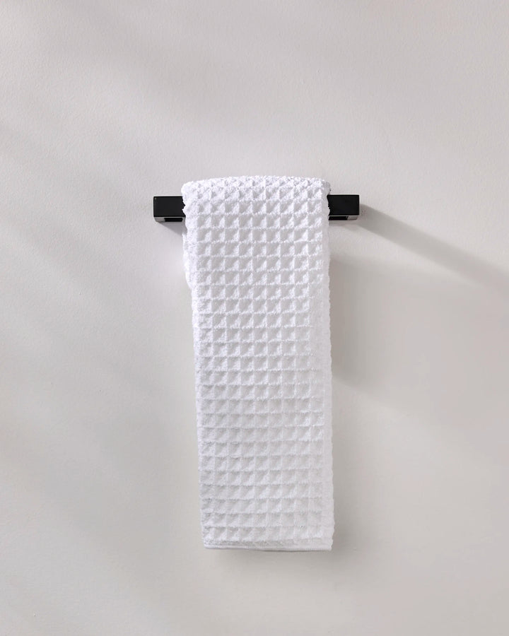 The GEOMETRY Cloud hand towel, made from recycled materials, in white waffle texture, hangs neatly on a black towel bar fixed to a light gray wall.