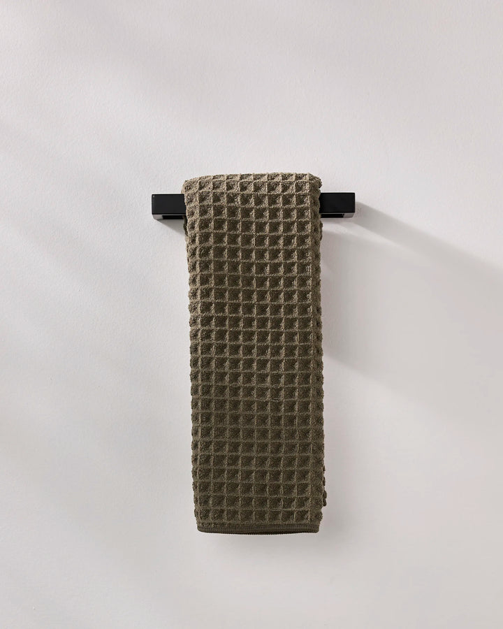 A GEOMETRY Forest ultra-absorbent waffle hand towel in dark gray hangs neatly on a black towel bar against a plain, light-colored wall.
