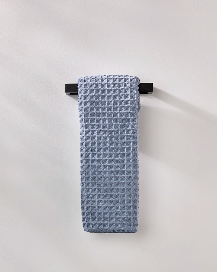 The GEOMETRY Sky hand towel, neatly folded in light blue waffle texture, hangs on a sleek black bar against a plain wall for an understated style touch.