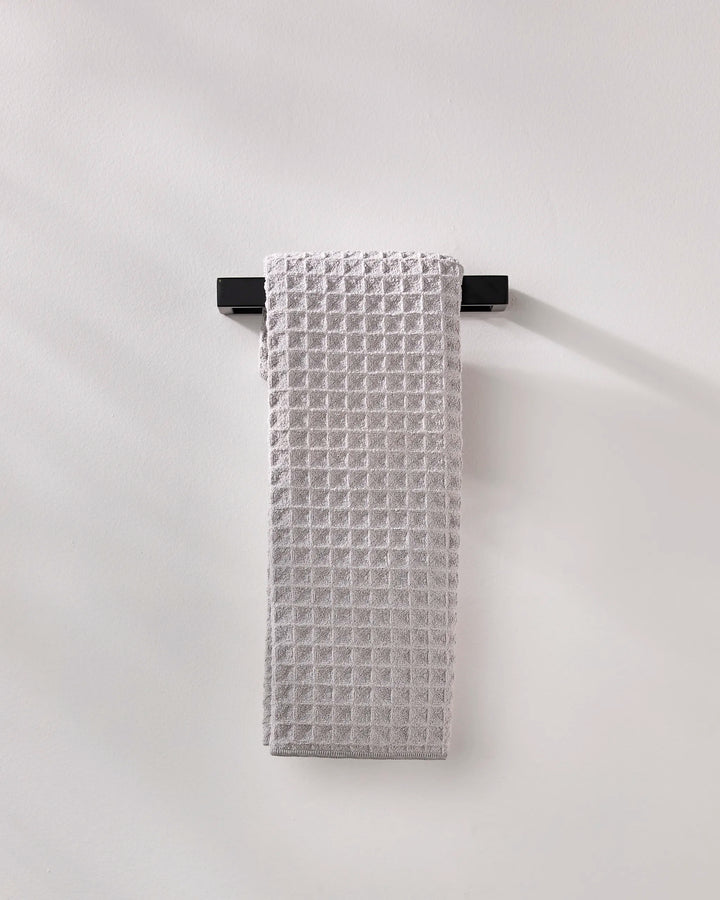 A light gray, waffle-textured GEOMETRY "Stone" luxury bath towel is neatly draped over a sleek black towel rack mounted on a plain, light-colored wall.