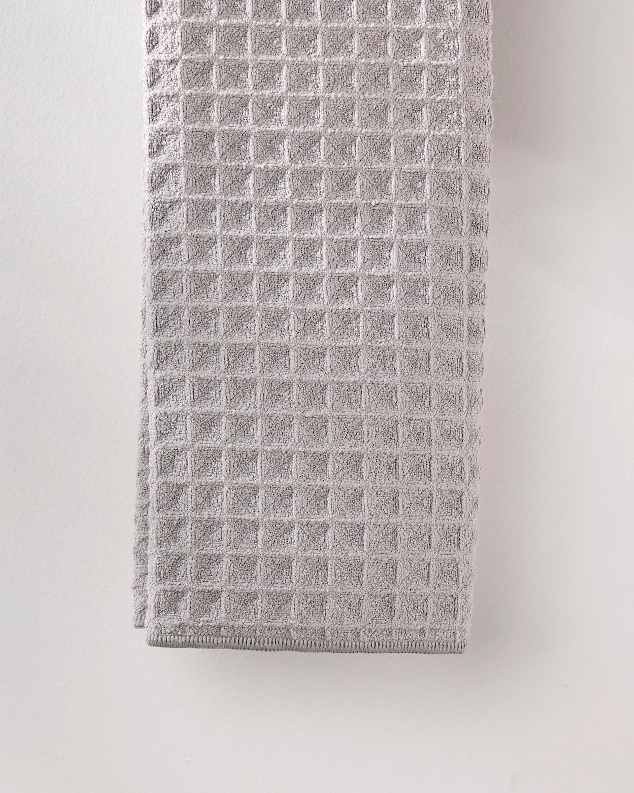 A GEOMETRY Stone hand towel in light gray, featuring a textured waffle pattern, hangs against a pale wall.