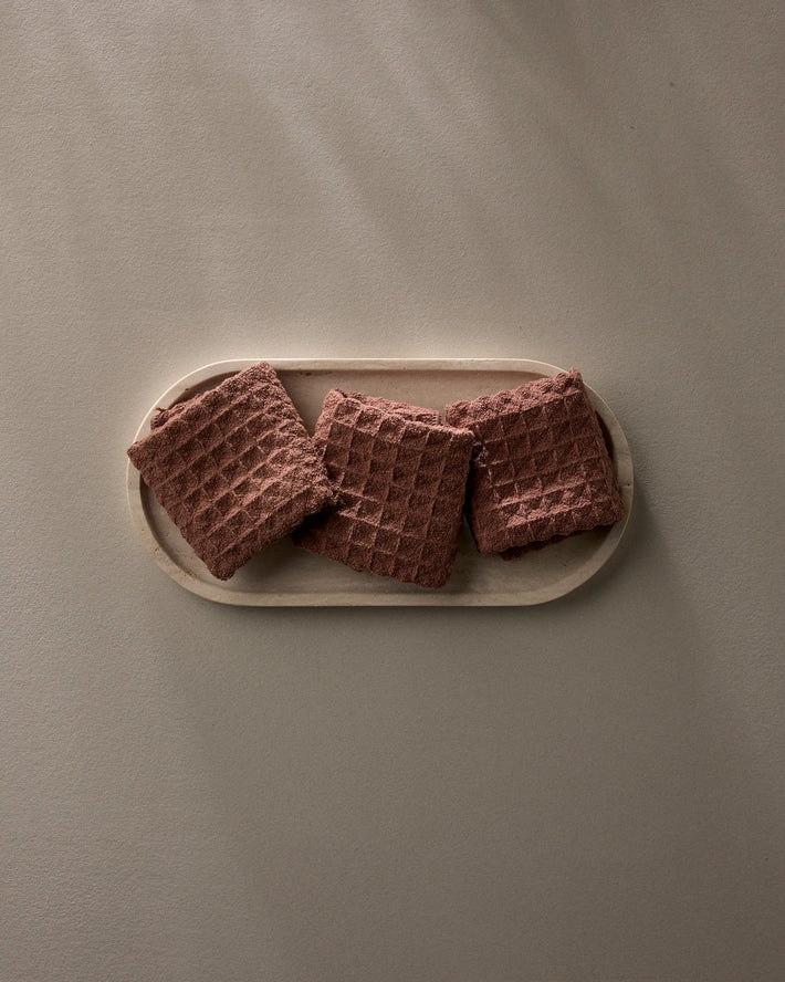 A wooden tray holds three folded, textured Dusty Rose washcloths by GEOMETRY on a smooth light beige surface with gentle natural light and soft shadows.
