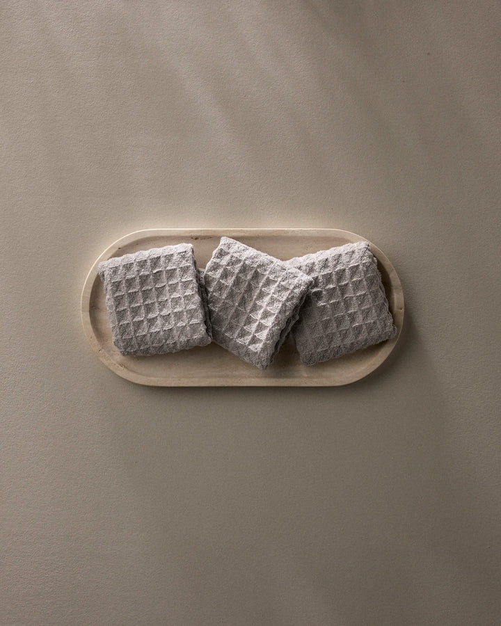 A wooden tray displays three neatly folded GEOMETRY Stone gray waffle washcloths, arranged in a row against a plain light beige background.