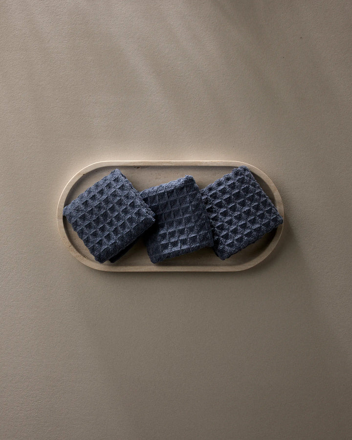 Three "Midnight Blue" waffle washcloths by GEOMETRY, made from recycled materials, are neatly displayed on a light oval tray atop a smooth taupe surface with gentle shadows.