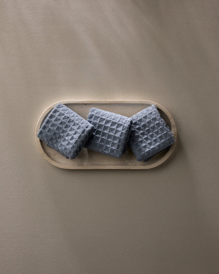 Three Sky washcloths by GEOMETRY, in gray waffle texture, are neatly folded side by side on a light wooden tray against a plain taupe background, bringing stylish luxury to your daily routine.