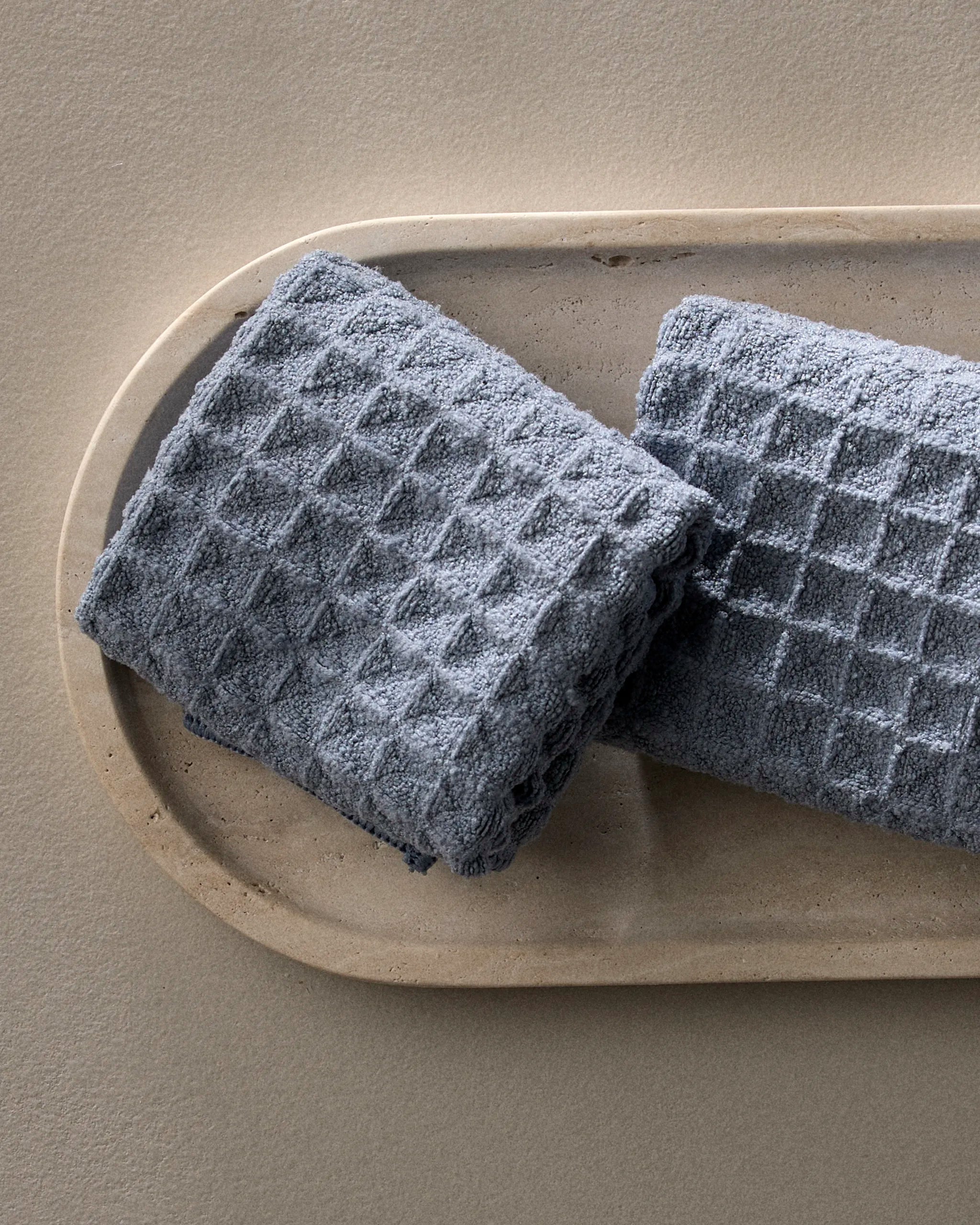 Two neatly folded, textured gray waffle washcloths from GEOMETRY's Sky collection rest on a beige oval tray against a neutral background, adding a touch of luxury to your space.