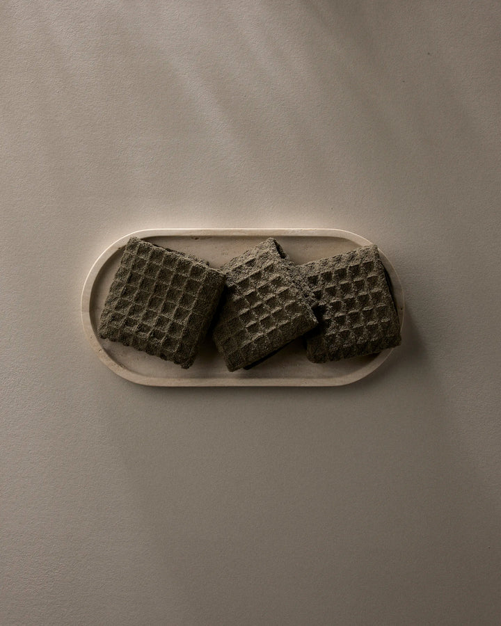 A beige oval tray displays three GEOMETRY Forest waffle washcloths in a dark, textured square design, set against a plain neutral background with soft lighting and gentle shadows.