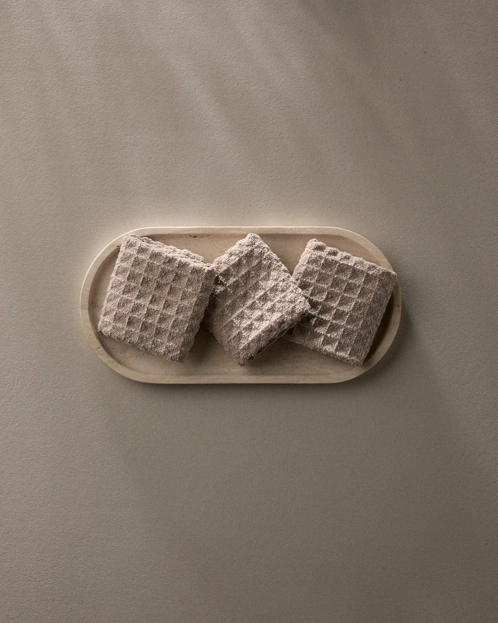 A Taupe oval plate by GEOMETRY displays three square, textured crackers with a waffle pattern—arranged side by side—on a neutral surface.