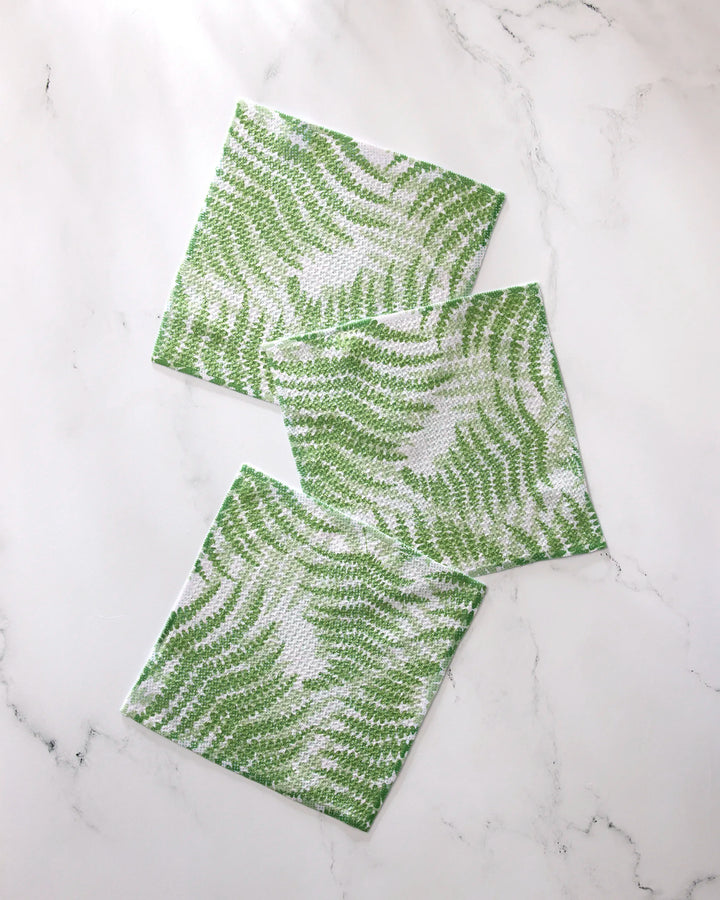Three square green and white GEOMETRY Swaying Leaf dishcloths, featuring a delicate wavy leaf pattern, are arranged in a staggered line on a white marble surface.