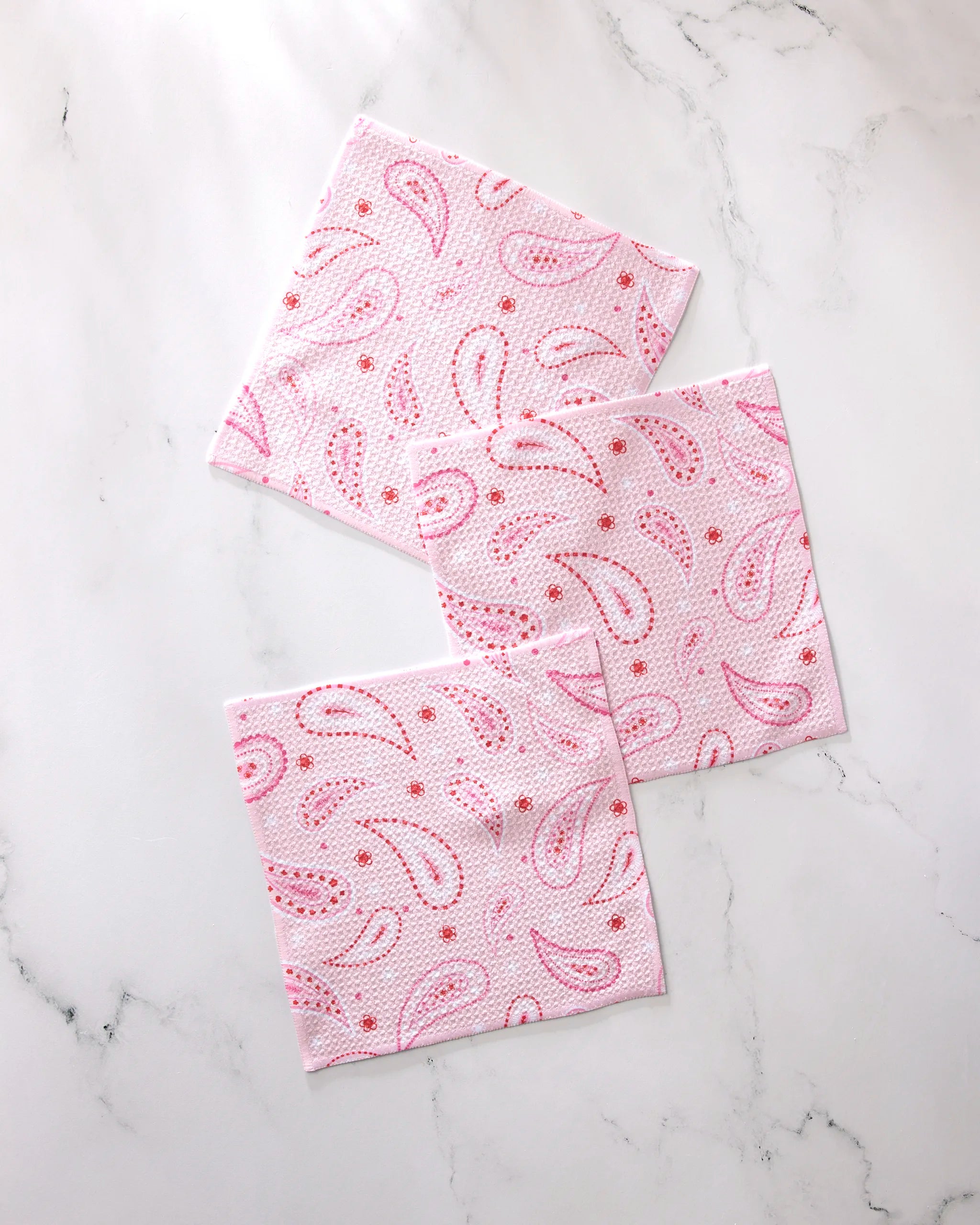 Three 'Sweet Paisley' napkins by GEOMETRY, featuring a pink paisley pattern and made from recycled materials, are displayed on a white marble surface.