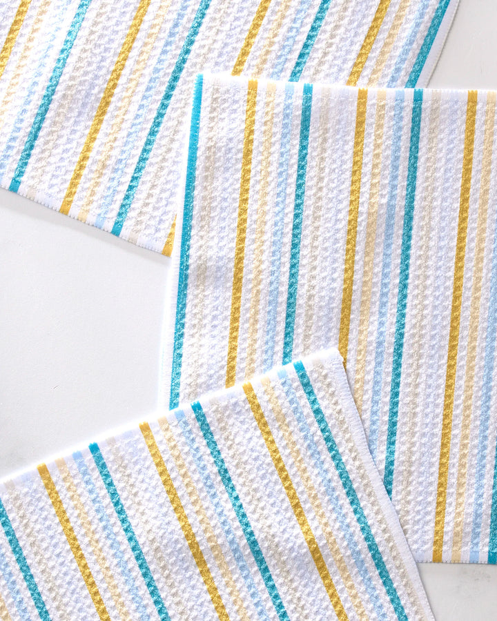 Three GEOMETRY Seaside Sunshine white textured recycled dishcloths with yellow, teal, and gray diagonal stripes are partially overlapping on a white surface.
