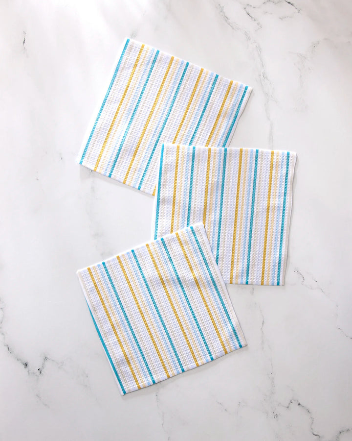 Three folded GEOMETRY Seaside Sunshine kitchen towels with blue, yellow, and white stripes are displayed on a white marble surface.