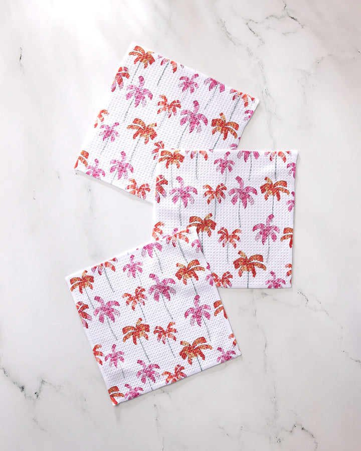 The GEOMETRY Pink Palms dishcloth set includes three super-absorbent towels, each adorned with pink and orange palm tree patterns on a white marble background.