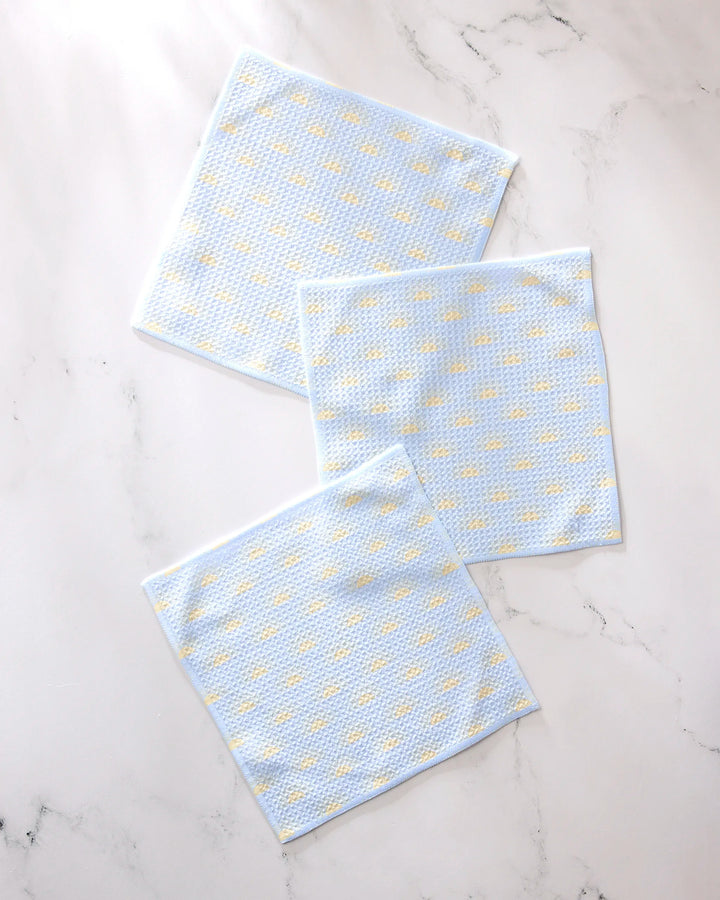 Three GEOMETRY "Soft Summer Sun" light blue textured recycled dishcloths with small yellow dots are arranged on a white marble surface.