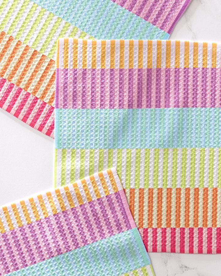Three pastel-striped Neon Nights dishtowels by GEOMETRY in purple, orange, yellow, green, and blue are arranged on a white surface with overlapping corners.