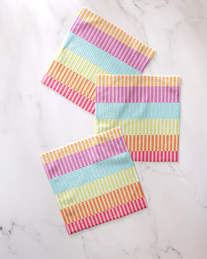 Three square GEOMETRY Neon Nights dishcloths with vibrant horizontal stripes in yellow, pink, green, blue, and purple are displayed on white marble—crafted from recycled materials.
