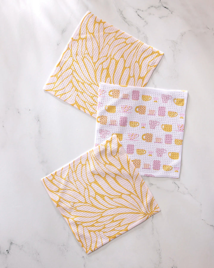 The GEOMETRY "Tenderhearted" set features three white dishcloths—two with golden yellow leaf patterns and one with a pink and yellow abstract geometric design—all made from recycled materials.