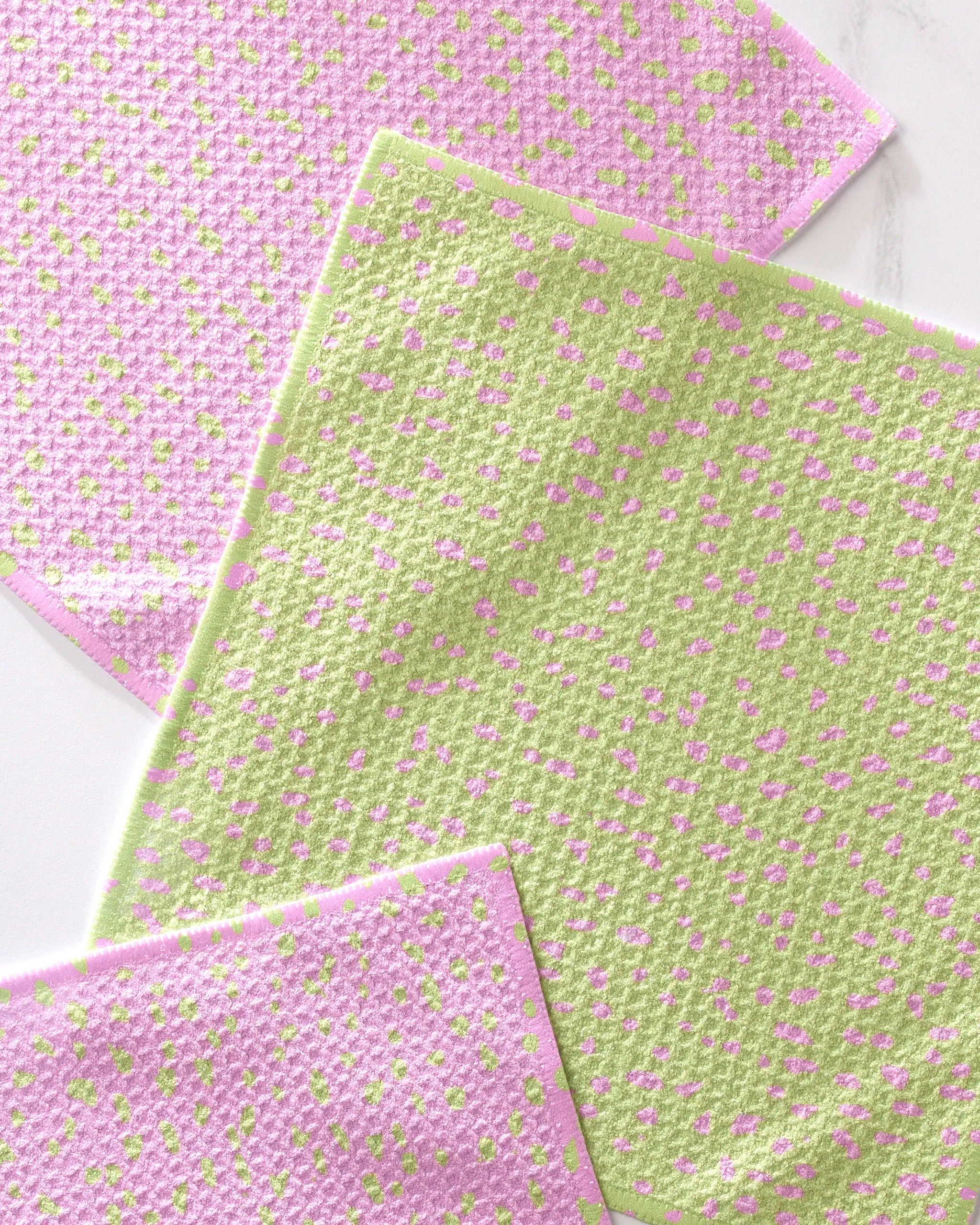 Three Speckle Green Pink cleaning cloths by GEOMETRY, featuring two pink with green dots and one green with pink dots, overlap stylishly on a light surface—perfect pastels for your dishcloth set.