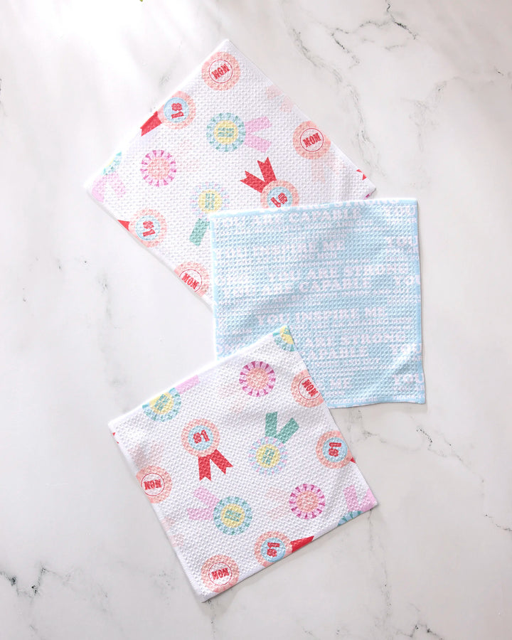 Three GEOMETRY Mom Mantra Swedish dishcloths, made from recycled materials, rest on marble; two showcase ribbon and medal patterns, while one is light blue with white text—an eco-friendly, super-absorbent kitchen set.