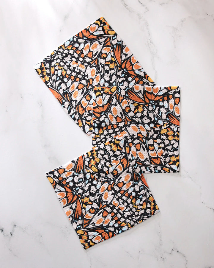 Three "Wings in the Wind" fabric napkins by GEOMETRY, featuring a vibrant monarch butterfly wing pattern in orange, black, yellow, and white recycled material, are arranged in a scattered layout on a white marble surface.