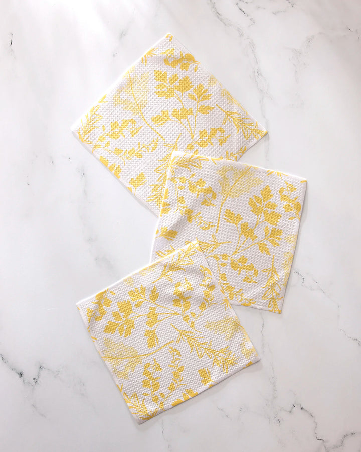 Three "Garden of Gold" cloth napkins by GEOMETRY, featuring a yellow leaf pattern on white and made from recycled materials, are arranged on a white marble surface.