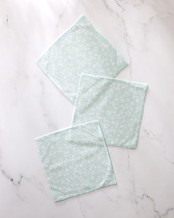 Three Dandy reusable dishcloths by GEOMETRY, featuring a light green and white textured design, are spread out on a white marble surface with their edges slightly overlapping.