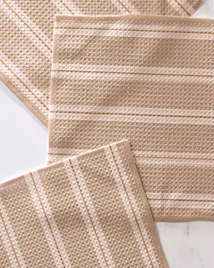 Three Vanilla Chai beige textured placemats by GEOMETRY, featuring subtle light stripes, are arranged partially overlapping on a white surface.