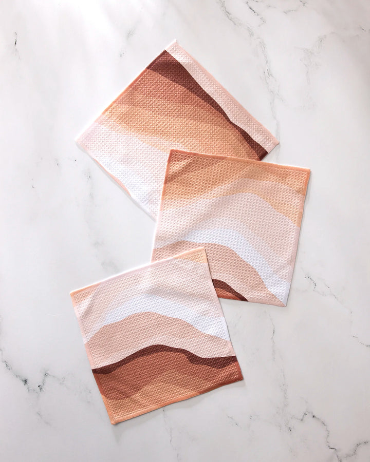 Three GEOMETRY "Levels of Fall" cloths, made from recycled materials and featuring abstract wavy patterns in brown, beige, and pink tones, are displayed on a white marble surface.