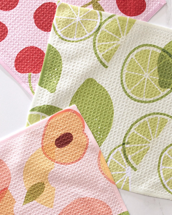 The GEOMETRY Fruit Fun dishcloth set includes three super-absorbent cloths featuring green limes, red cherries on pink, and playful peaches, arranged together for a practical and cheerful kitchen accent.