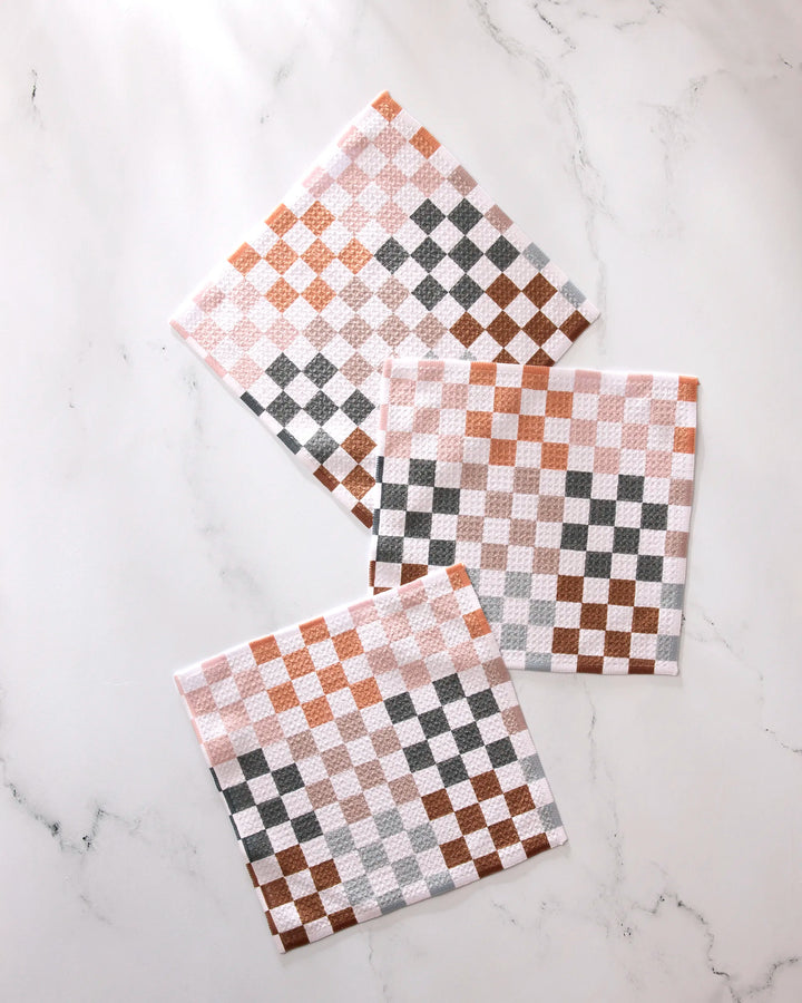 Three GEOMETRY Autumn Checkers square napkins in white, brown, and gray checkered patterns are arranged in a casual, overlapping layout on a white marble surface. This set features absorbent kitchen towels.