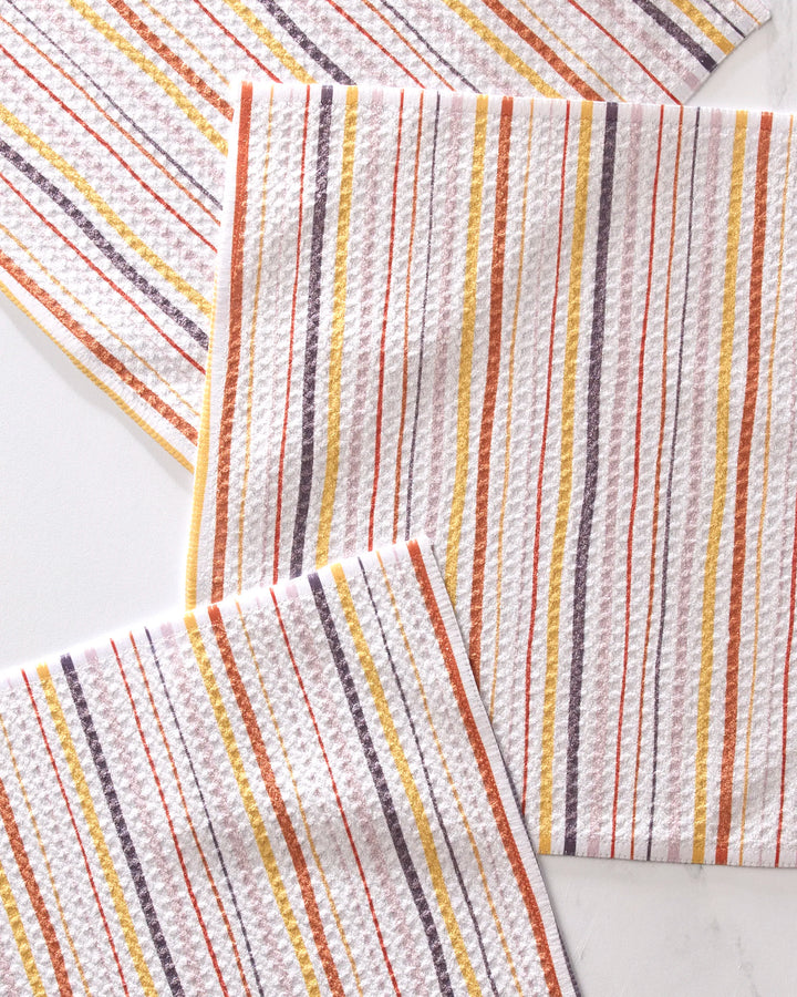 The GEOMETRY Stripe Season set includes three super-absorbent, textured white kitchen towels featuring vertical stripes in orange, yellow, red, and gray, stylishly arranged overlapping on a light background.